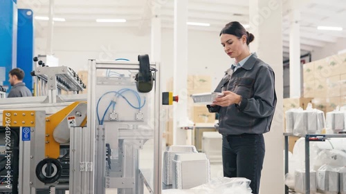 Female manager in uniform inspecting stack of foil containers in factory. Colleague working in background. Industrial production line.