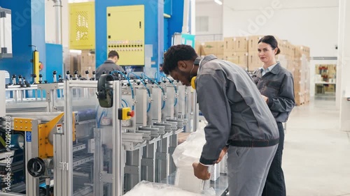 African American male manager wearing protective headphones while inspecting stack of foil containers in factory. Pointing to colleague about machine settings.