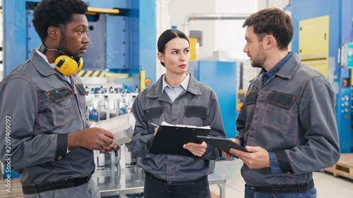 Group of factory engineers discussing project. Female holding blank showing data while colleague using tablet. Team collaborating in production facility.