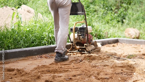 Fotografie Worker legs in protective boots stand beside compacting machine on sandy constru