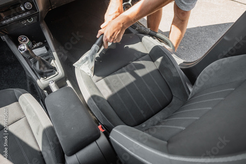 Person vacuuming car seat upholstery with hand nozzle in bright daylight, cleaning grey fabric interior, center console and stitching visible during detailing work