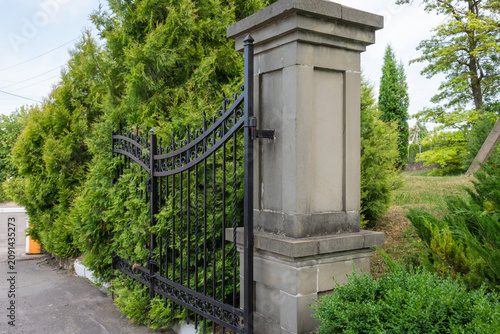 Принти на полотні A sturdy black metal gate is attached to a solid, light-colored stone pillar, surrounded by lush green coniferous trees