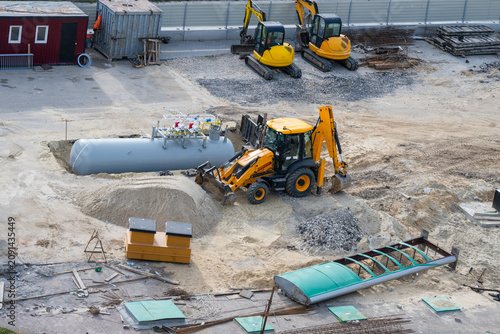Картината върху платно an active construction site features a yellow backhoe loader operating next to a large underground storage tank, with two mini-excavators parked in the background