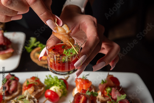 Photos Close-up of a woman's hands with long manicured nails holding a small glass of gazpacho cocktail shooters with a shrimp skewer over a catering table with various appetizers