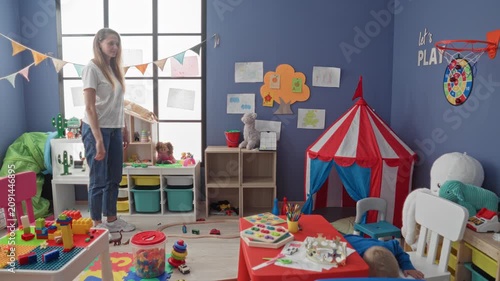 Woman holding toy car points to boy at cluttered red table in playroom building; parental care tenderness.