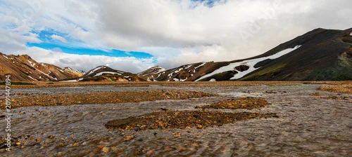 Panel kuchenny z motywem Braided glacial river winding through colorful rhyolite mountains with snow patches in Landmannalaugar, Iceland Highlands beneath dramatic clouds and vibrant evening light.