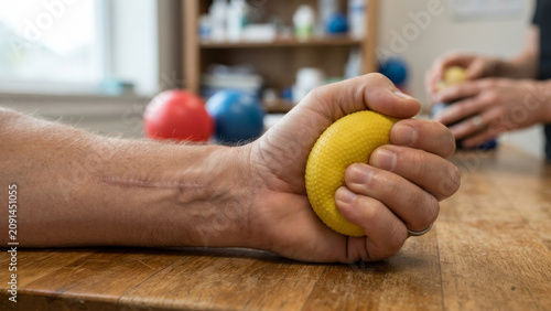 Close-up of a man's hand with a surgical scar squeezing a textured yellow therapy ball on a wooden table under soft natural light