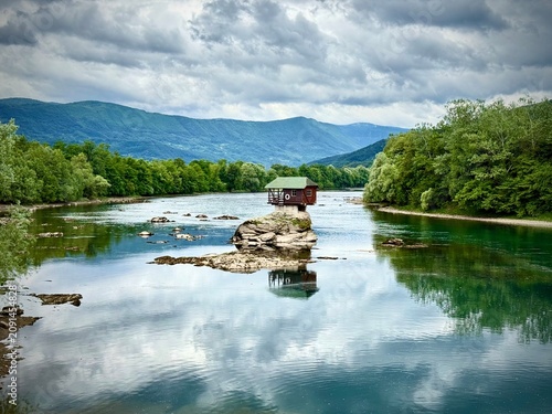 A small wooden house sits atop a rock in the middle of the Drina River in Serbia. Surrounded by lush green forests and mountainous terrain, the house appears isolated and tranquil. The water is calm