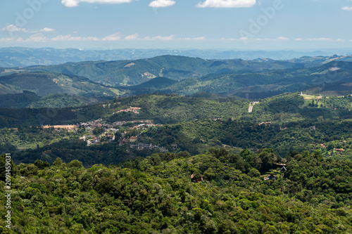 Panoramic view of Monte Verde valleys with dense forest, rolling mountains and small town under summer morning sunny clouded blue sky. Monte Verde, Camanducaia, Minas Gerais - Brazil.