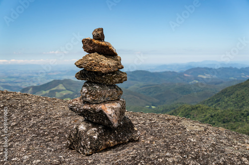 Balanced stone stack on mountain viewpoint overlooking vast green valleys under summer morning sunny clear bllue sky in Monte Verde, Camanducaia, Minas Gerais - Brazil