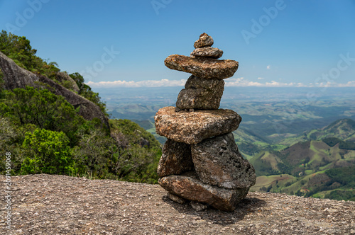 Balanced stone stack overlooking vast green valleys saw from Pedra Redonda rock formation summit under summer morning sunny clear blue sky in Monte Verde, Camanducaia, Minas Gerais - Brazil.