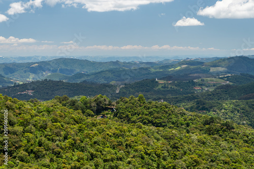 Panoramic Atlantic forest valleys and rolling hills beneath some clouds saw from Pedra Redonda rock formation summit under summer morning blue sky in Monte Verde, Camanducaia, Minas Gerais, Brazil