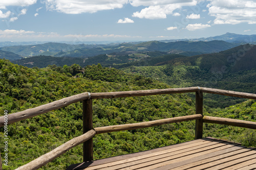 Panoramic view from wooden observation deck lookout over lush green mountains and Atlantic Forest under blue sky with scattered clouds in Monte Verde, Camanducaia, Minas Gerais - Brazil.