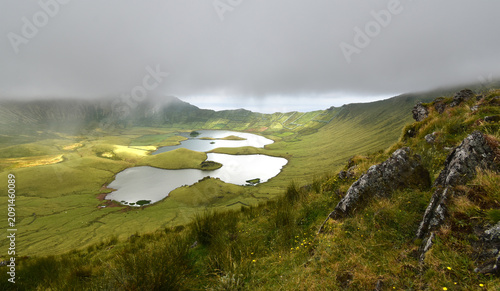 Canvastavla A breathtaking panoramic view of the Caldeirão caldera with its tranquil lakes, surrounded by lush green terraced slopes and dramatic rock formations under a cloudy sky, Corvo Island, Azores