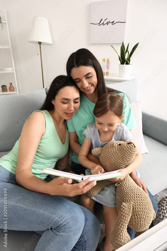 Obraz premium Young lesbian couple with adopted little girl reading book at home