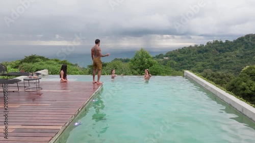 Group of young people dancing by pool at small party in tropical resort. Tattooed man stands on wooden deck while women swim and socialize in turquoise infinity pool overlooking lush jungle and ocean.