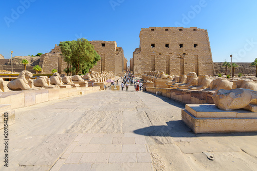 Tourists on The Way of the Rams featuring ram headed sphinxes lined on both sides as they approach the First Pylon of the Karnak Temple Complex in Luxor Egypt.