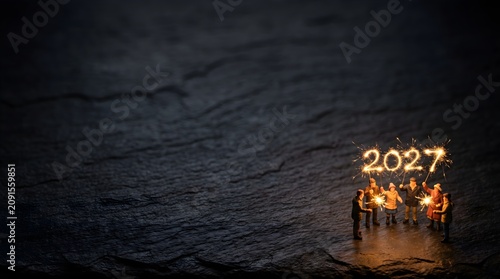 Miniature people celebrating new year 2027 with glowing sparkler numbers on dark slate background