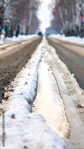 A wintry road with snow, mud, and tire tracks. A car appears at a distant horizon on a long stretch. The foreground is wet