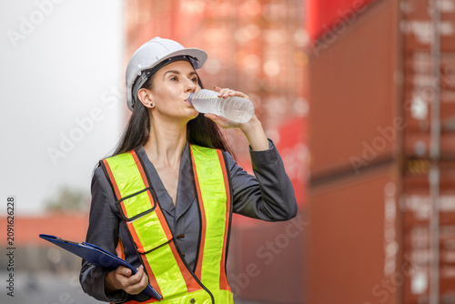 Female engineer drinking water during a break at cargo shipping port, Woman wearing safety gear holding a clipboard and drinking water at a shipping container terminal
