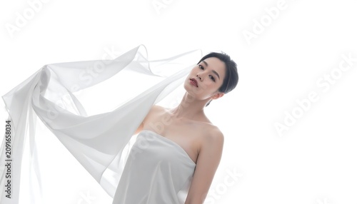 A woman draped in white fabric poses against a white background. Her head is tilted, and she gazes upward
