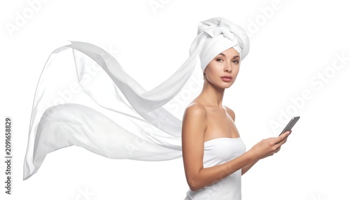 A woman, draped in white towel, stands against white background, holding a phone. Long flowing fabric adds movement and elegance