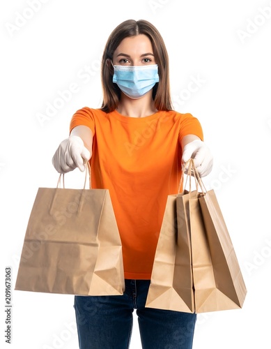 A woman in an orange shirt, mask, and gloves extends two shopping bags towards the viewer, isolated on white