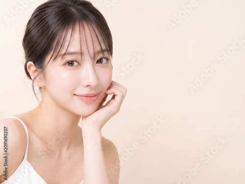 淡いベージュ背景で頬に手を添えて自然に微笑む若い日本人女性、明るくやさしい印象のスキンケア・美容広告ポートレート