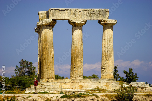 A man in a red sweater and tan pants stands near the ancient ruins of the Temple of Apollo in Corinth, Greece, under a clear blue sky.