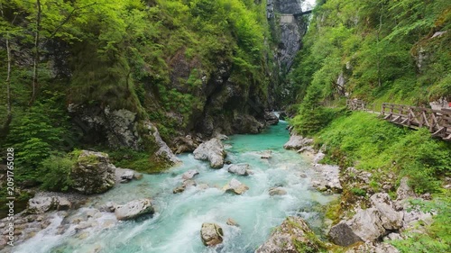 Turquoise water rushes through Tolmin Gorge surrounded by mossy rocks and vibrant green forest with a wooden walkway along the riverbank.