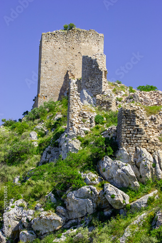Ruins of a stone tower on a rocky, overgrown hillside under a clear blue sky.