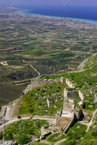 A high-angle view of the ancient ruins of Acrocorinth, a fortress overlooking the modern city of Corinth and the Gulf of Corinth in Greece. 