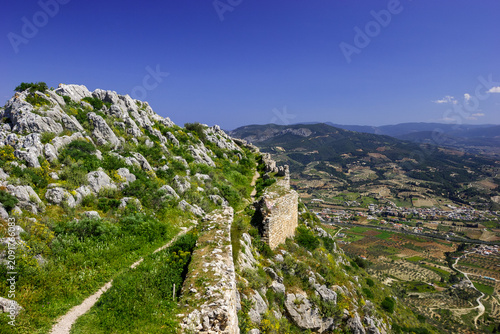 A scenic view of the ancient ruins of a castle on a rocky, green mountainside overlooking a valley with a village and agricultural fields under a clear blue sky.