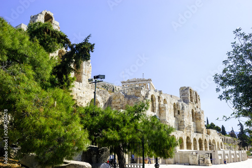 The Odeon of Herodes Atticus, a stone theatre structure on the Acropolis of Athens, Greece, on a clear day with lush green trees in the foreground.