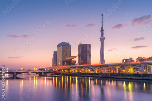 a vibrant cityscape at dusk, featuring modern skyscrapers on the Sumida River. reflecting in the calm river below. The sky is painted with soft hues of purple and orange.