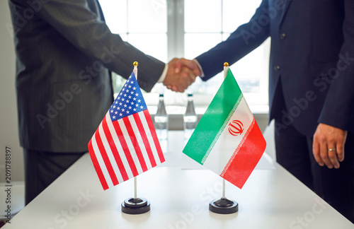 Iran USA flags handshake diplomacy agreement negotiation. Two suited diplomats shake hands at a table as flags signal politics and treaty talks. Strong symbol of international diplomatic accord.