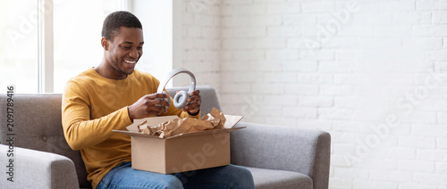 Happy man unboxing package on sofa, holding new white wireless headphones in a bright modern living room with a white brick wall, ideal for online shopping delivery concepts