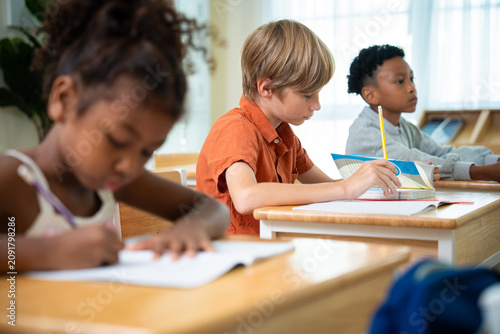Focused elementary schoolboy reading a textbook and writing at his classroom desk. Concentrated primary student studying and turning pages of a book during a class lesson.
