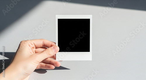 Hand holding blank instant photo frame mockup with black space on grey background with sunlight and shadows.