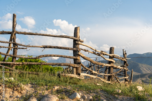 An old, weathered wooden fence made of rough tree branches and logs stands on a grassy, rocky hill.