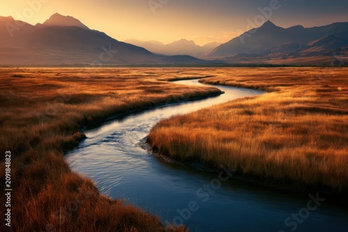 Sinuous stream curving across golden grassland plains under a warm sunset sky with jagged peaks for wilderness conservation and travel marketing