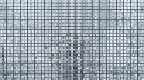 Textured mosaic glass panel with blurred human silhouette behind grid pattern in interior setting, translucent frosted squares diffusing light and providing privacy and abstraction