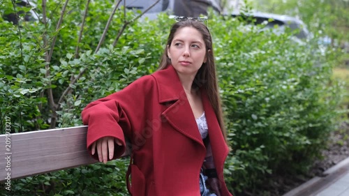 A beautiful young woman with long hair, wearing a red coat, sits on a bench daydreaming on a spring day.