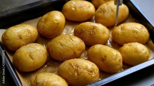 Roasted whole golden potatoes on baking tray lined with parchment paper being seasoned with olive oil, close up view of homemade oven cooked side dish
