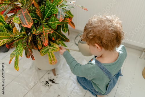 A curious toddler examines a potted plant and croton with a trowel on a bright white floor. The wondering child touches leaves and foliage discovering indoor greenery in a calm organized home.