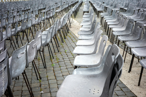 Фотография A vast expanse of empty grey plastic chairs is arranged in precise, repetitive rows across a rugged, weathered stone-paved public square