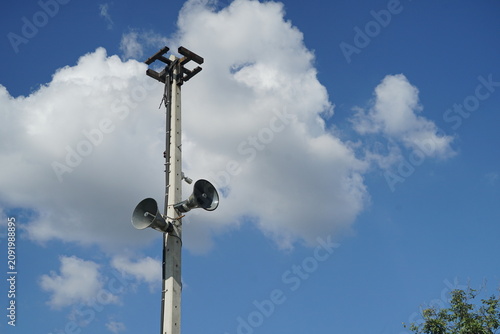 Outdoor loudspeakers mounted on tall metal pole against blue sky with clouds