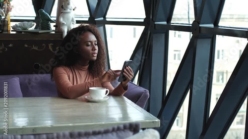 Cute African American woman drinking coffee or tea and using mobile phone in cafe