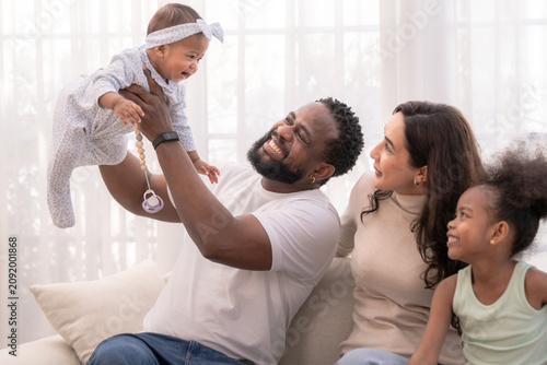 Fotografie A fun-loving father with his baby holds up his giggling baby to play on a sunlit white sofa, while the daughter cheers happily full of natural candid warmth captured in soft natural light