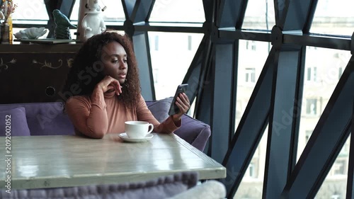Cute African American woman drinking coffee or tea and using mobile phone in cafe